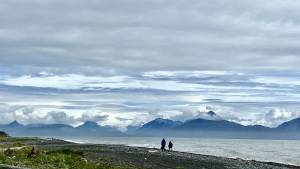 Two people walk along a beach, with the waters of Kachemak Bay behind them, with views of jagged mountains and glaciers covered by a partly cloudy blue and gray sky.